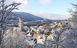 Verschneite Stadt mit Kirchturm und Hügeln im Hintergrund.