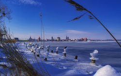 Winterliches Ufer mit gefrorenem Wasser und Stadt im Hintergrund, blauer Himmel