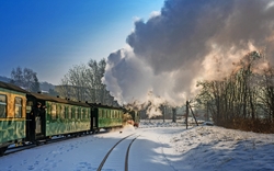 Grüner Dampflokzug auf verschneiter Strecke, Rauch steigt empor, Bäume im Hintergrund.