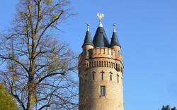 Turm mit spitzen Dächern neben einem Baum unter blauem Himmel.