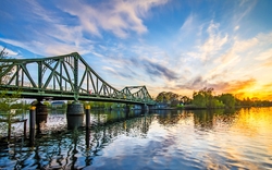 Brücke bei Sonnenuntergang über einem Fluss mit spiegelnder Wasseroberfläche und blauem Himmel.