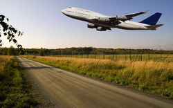 Flugzeug fliegt über ländliche Straße und Wiese bei klarem Himmel.