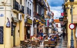 Menschen sitzen an Straßencafé-Tischen einer belebten, bunten Gasse.