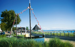 Boot mit Fahnenmast am Strand mit Promenade und See im Hintergrund.