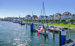Hafen mit Segelbooten und Häusern im Hintergrund bei sonnigem Wetter.