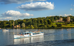 Ausflugsboot auf einem Fluss vor einer malerischen Landschaft mit Schloss im Hintergrund.