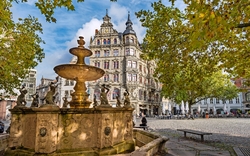 Historischer Brunnen und Gebäude an einem sonnigen Platz, umgeben von Herbstbäumen.
