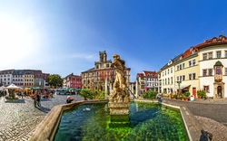 Marktplatz mit Brunnen und historischen Gebäuden bei Sonnenlicht.