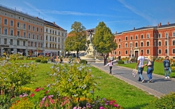 Stadtplatz mit Blumen, Springbrunnen und Personen bei sonnigem Wetter.