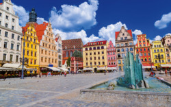 Marktplatz mit bunten Gebäuden und Springbrunnen unter blauem Himmel.