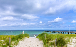 Sandpfad mit Gras führt zum Strand und Meer, seitlich Steg unter bewölktem Himmel.