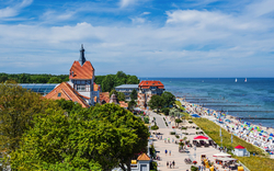 Küste mit Promenade und Gebäuden, im Hintergrund das Meer und Segelboote.