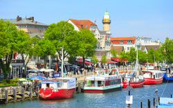 Hafen mit Booten und historischen Gebäuden, Leuchtturm im Hintergrund.