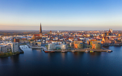Luftaufnahme einer Stadt mit Wasser, modernen Gebäuden und einer Kirche am Horizont.