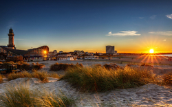 Sonnenuntergang am Strand mit Dünen und Leuchtturm im Hintergrund.