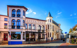 Historisches Gebäude mit Türmchen und Arkaden in einer sonnigen Straßenszene.