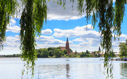 Blick auf eine Kirche und See, umrahmt von herabhängenden Zweigen.