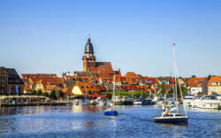 Stadtansicht mit Kirchturm und Segelbooten auf einem Fluss.