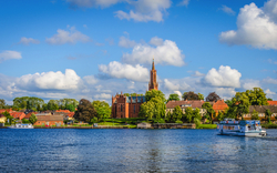 Idyllische Uferlandschaft mit Kirche und Boot auf einem Fluss bei blauem Himmel.