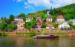 Schiff auf dem Neckar in Heidelberg, im Hintergrund historische Gebäude und bewaldete Hügel.