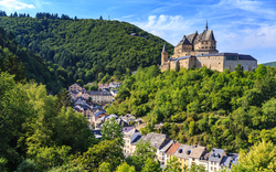 Burg auf Hügel mit grünen Wäldern und Dorf im Tal bei sonnigem Himmel.