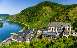 Mittelalterliches Dorf mit Kirche an einem Fluss in einer grünen Hügellandschaft.