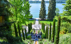 Gartenanlage mit hohen Zypressen und Blick auf einen See im Hintergrund.