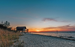 Sonnenuntergang am Seeufer mit einem hölzernen Gebäude auf Pfählen und Kiesstrand im Vordergrund.