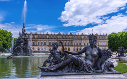Brunnen mit Skulpturen vor einem Schlossgebäude bei sonnigem Wetter.