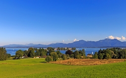 Blick auf eine grüne Landschaft mit See und Bergen im Hintergrund bei klarem Himmel.
