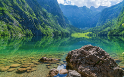 Bergsee mit klarem Wasser und Felsen im Vordergrund, umgeben von bewaldeten Bergen.