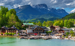 Idyllisches Dorf am See mit Bergen im Hintergrund bei bewölktem Himmel.