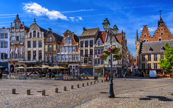 Marktplatz mit historischen Gebäuden, Cafés und blumengeschmückter Straßenlaterne bei Sonnenschein.