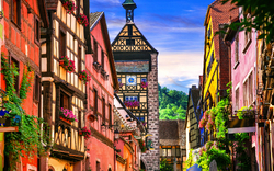 Bunte Fachwerkhäuser in einer engen, blumengeschmückten Gasse mit Turm im Hintergrund.