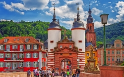 Stadttor auf einer Brücke in Heidelberg mit Menschen und Fachwerkhäusern im Hintergrund.