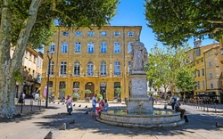 Platz mit Springbrunnen und Statue vor historischem Gebäude, Menschen entspannen im Schatten.