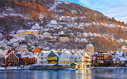 Winterlandschaft mit bunten Häusern am Wasser und schneebedeckten Hügeln im Hintergrund.