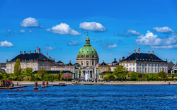 Kirche mit grünlicher Kuppel und Gebäuden am Wasser unter blauem Himmel.