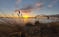 Sonnenuntergang über einer Brücke am Wasser mit Gras im Vordergrund.
