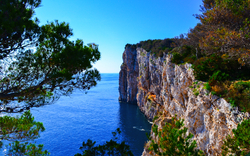 Klippen am Meer mit blauem Himmel und Bäumen im Vordergrund.
