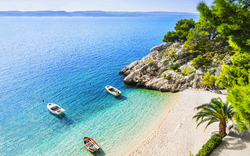 Strand mit klarblauem Wasser, Booten, Felsen, Bäumen und weißem Sand