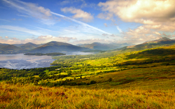 Hügelige Landschaft mit See und bewölktem Himmel im Hintergrund.