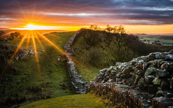 Sonnenaufgang über einer historischen Steinmauer in hügeliger Landschaft.