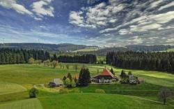 Landschaft mit Wiesen, Wald und einem einsamen Bauernhof unter blauem Himmel.