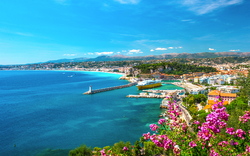 Blick auf Küstenstadt und Hafen am Mittelmeer mit blühenden Blumen im Vordergrund.