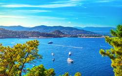 Küste mit blauem Meer, Segelbooten und bewaldeten Hügeln im Hintergrund.