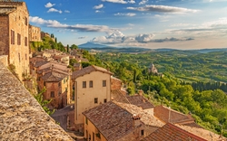 Hügelige Landschaft mit mittelalterlichen Gebäuden und bewaldeten Tälern bei Sonnenuntergang.