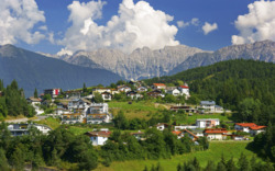 Häuser auf einem grünen Hügel mit Bergen und Wolken im Hintergrund.