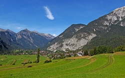 Blick auf ein Dorf in den Alpen, umgeben von Wiesen und Bergen unter blauem Himmel.
