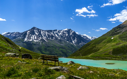 Bank vor Bergsee und schneebedeckten Bergen unter blauem Himmel.